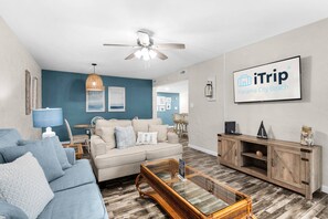A modern living room with a blue accent wall, a ceiling fan, a large TV displaying 'iTrip Panama City Beach', and a wooden cabinet. - Sugar Beach C-8 (Panama City Beach)