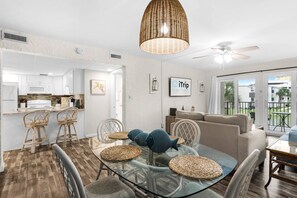 A modern kitchen with white cabinets, a microwave, and a stove. There is a dining area with a glass table and wicker chairs. A ceiling fan and pendant light are visible. - Sugar Beach C-8 (Panama City Beach)
