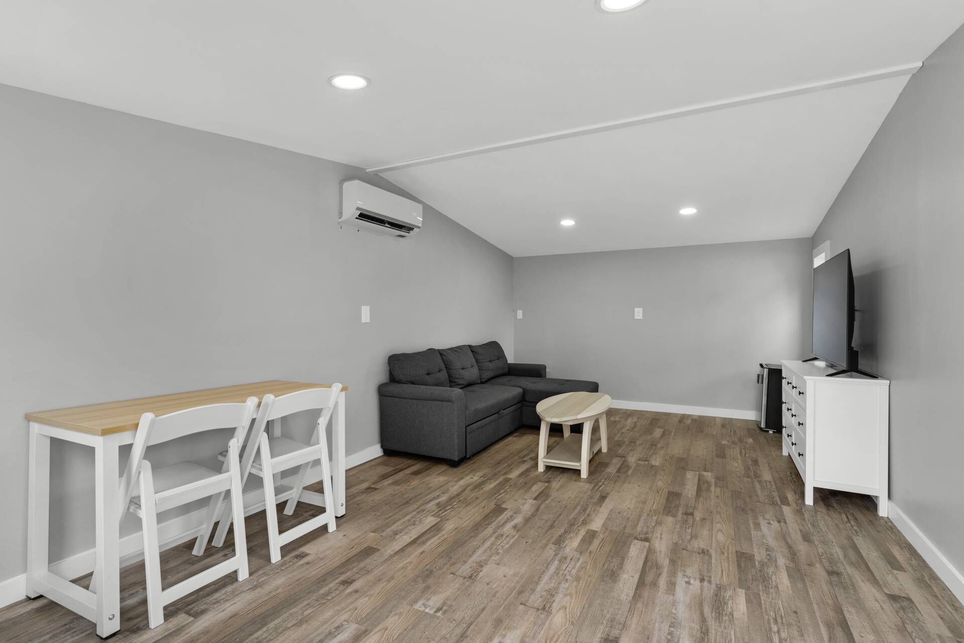 Un salon moderne avec un canapé gris, une table en bois avec des chaises, un téléviseur posé sur un meuble blanc et un appareil de climatisation fixé au plafond.