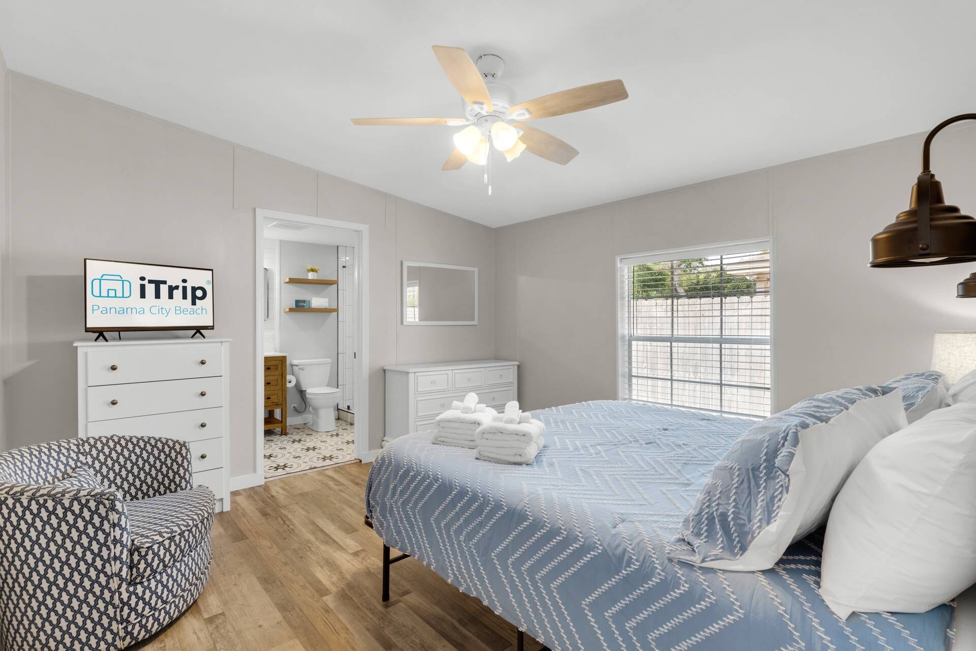 A bedroom with a bed, a ceiling fan, a TV, a window, and a bathroom visible through an open door.