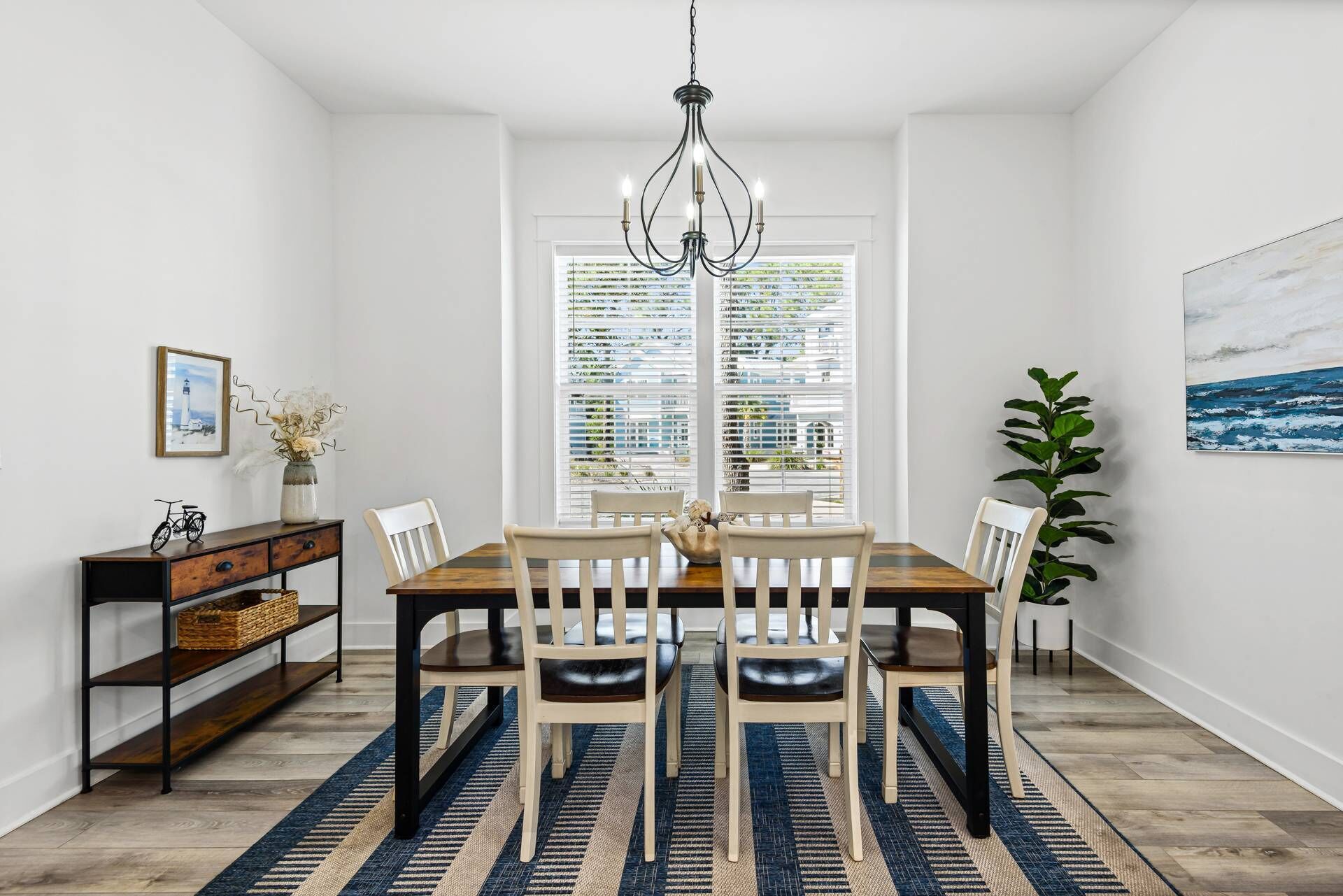 A dining area with a wooden table and chairs, a chandelier, a bookshelf, and a large window with blinds.