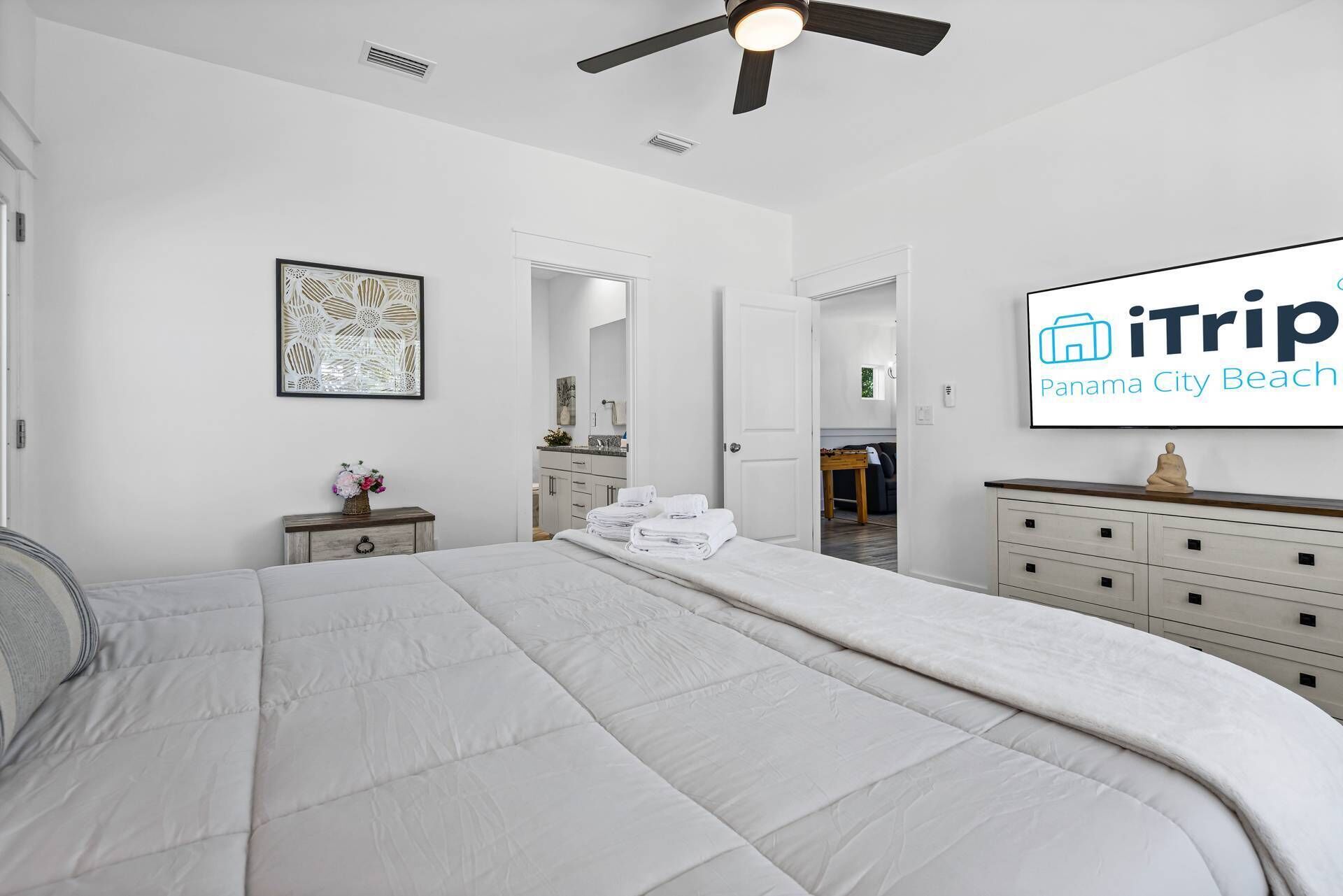 A hotel room with a large bed, a ceiling fan, a bathroom with a mirror, and a TV displaying 'iTrip Panama City Beach'.