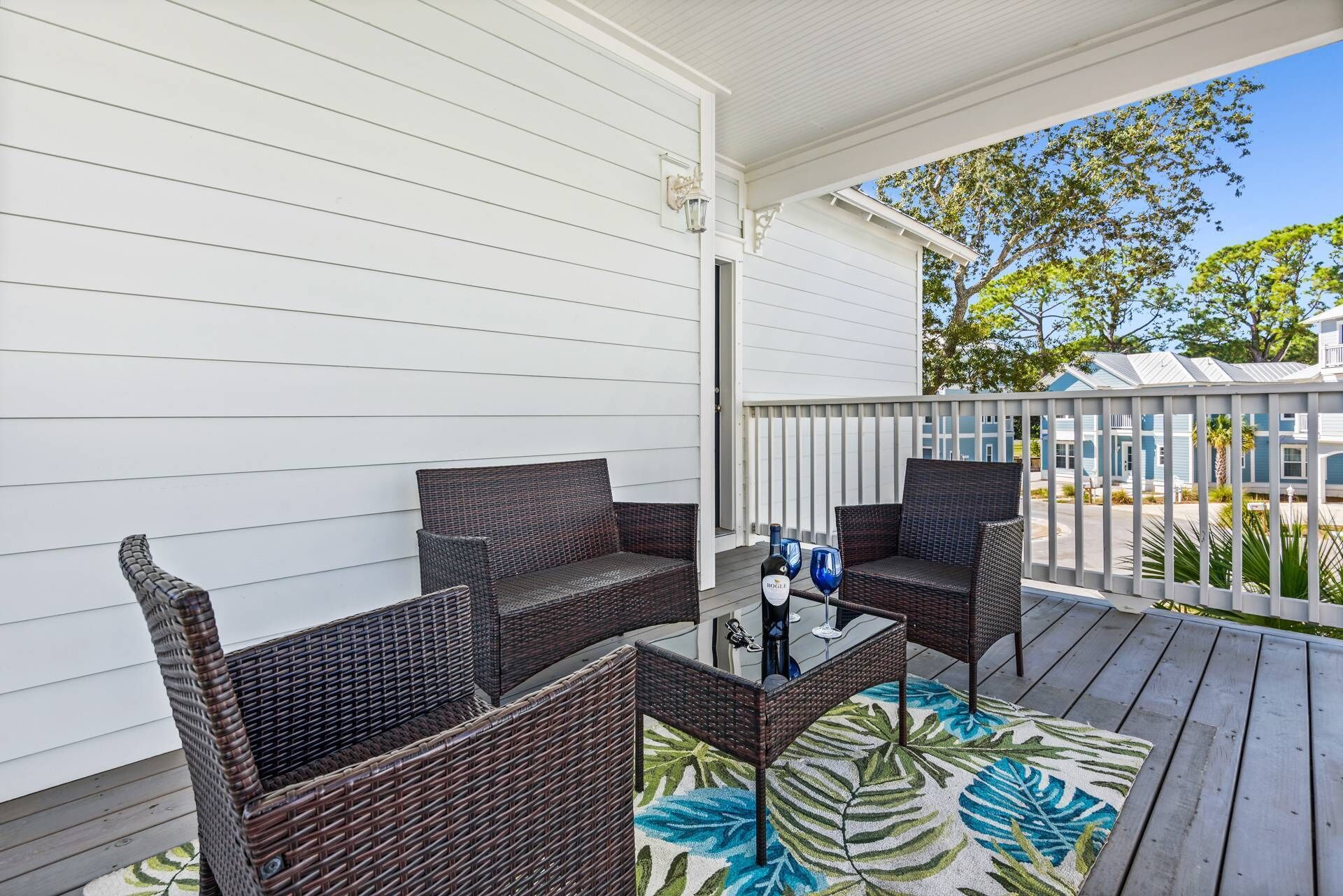 A covered outdoor seating area with wicker furniture and a glass table, featuring a tropical leaf-patterned rug.