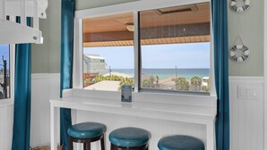 Quarto com uma grande janela com vista para a praia, uma mesa consola branca com três banquinhos e vista para o oceano.