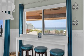 Ein Zimmer mit großem Fenster mit Blick auf einen Strand, ein weißer Konsolentisch mit drei Hocker und Meerblick.