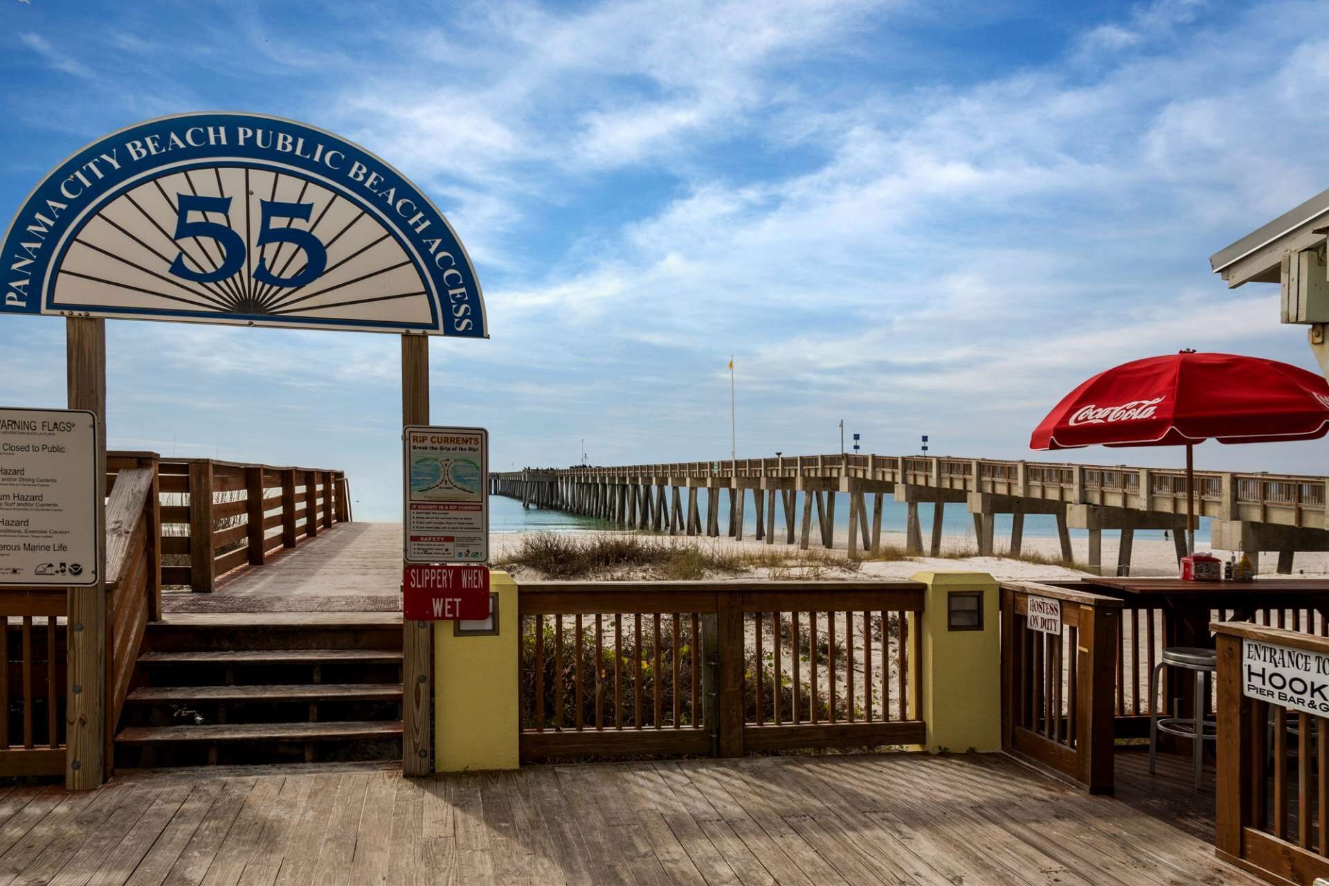 A wooden pier leading to a beach area with a Coca-Cola umbrella, warning signs, and a sign indicating the entrance to a pier bar and grill.