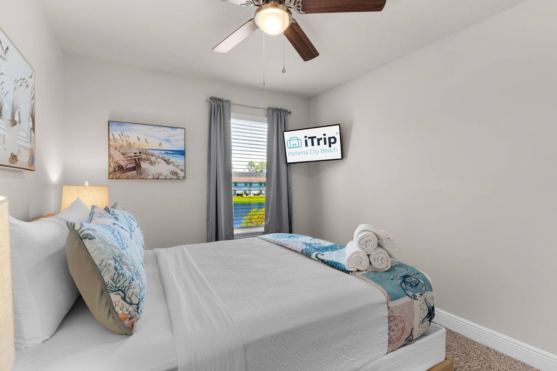 A bedroom with a bed, a ceiling fan, a TV displaying 'iTrip Panama City Beach', and a window with curtains.