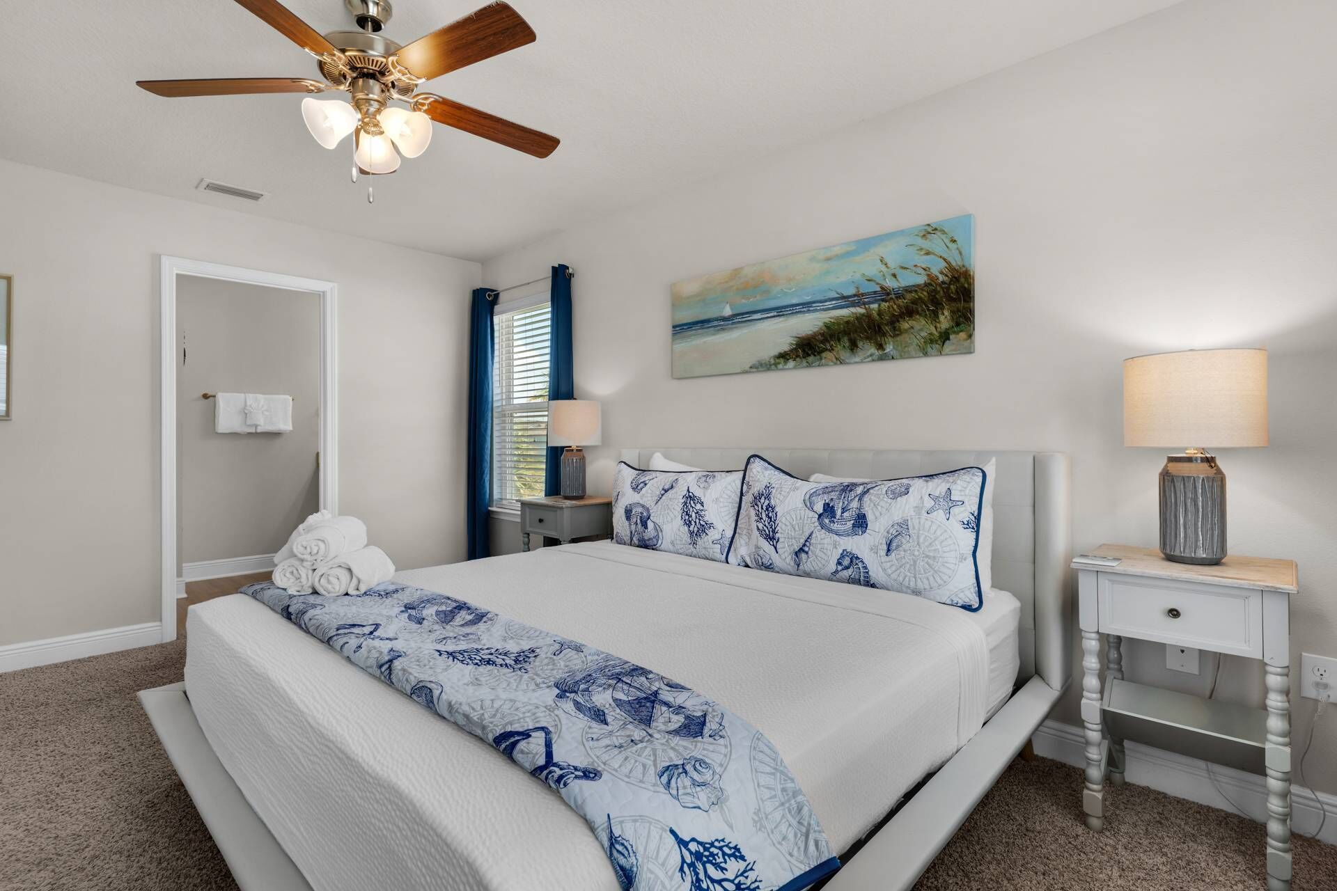 A bedroom with a bed, a ceiling fan, a painting, a nightstand, and a lamp.