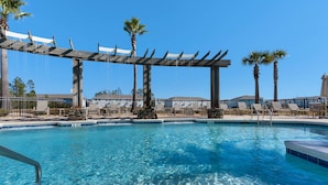 Piscina cristallina blu con una pergola in legno sopra, circondata da palme e poltroncine relax.