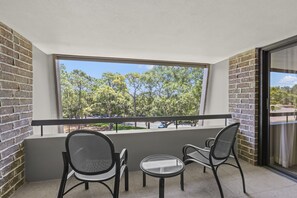 A balcony with two chairs and a small table, a large window with a view of trees, and brick walls.