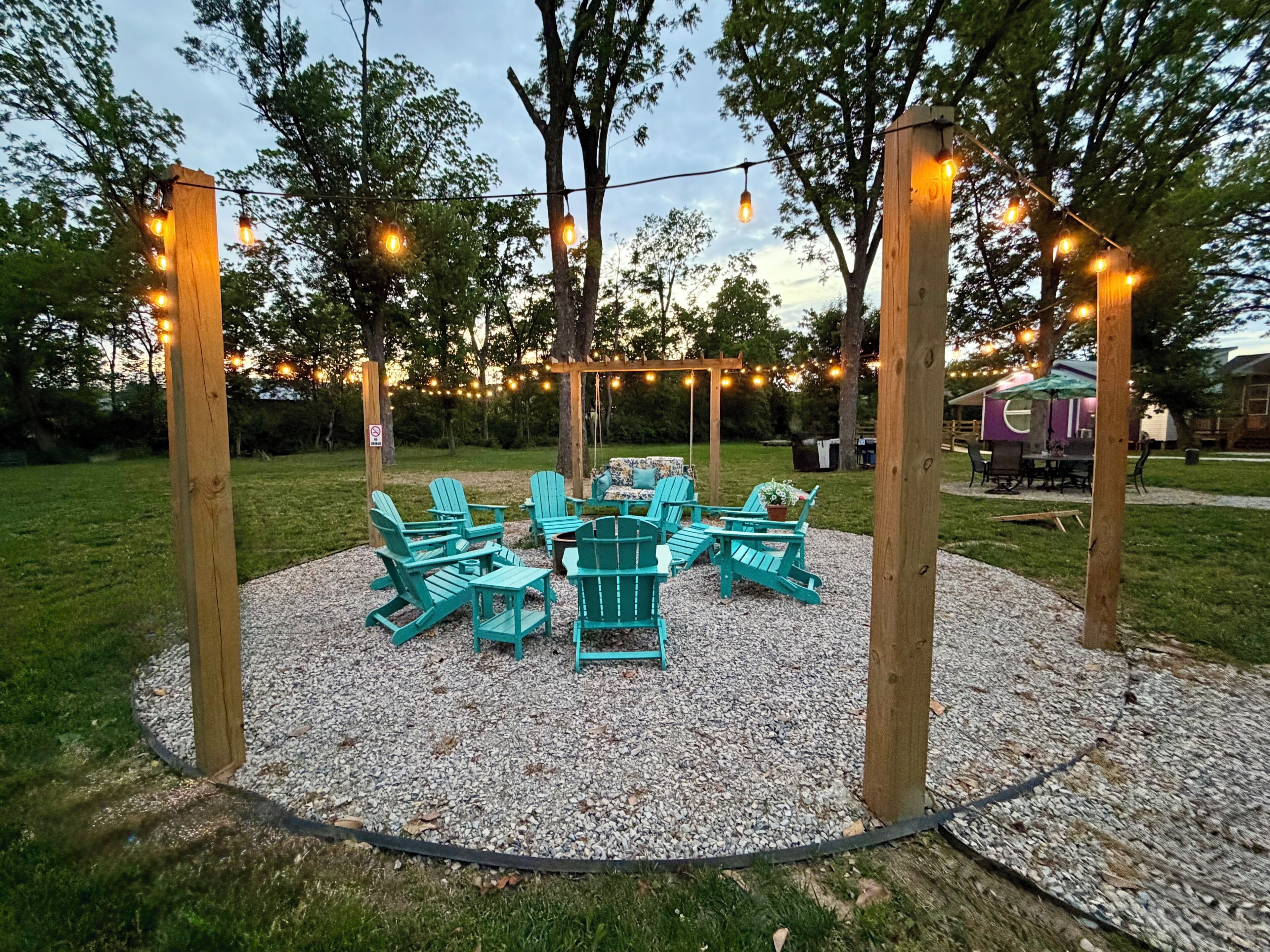 An outdoor seating area with Adirondack chairs and a wooden pergola, surrounded by trees and string lights.