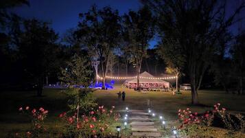 Ein nächtliches Freiluft-Event mit einem Zelt, das mit Lichterketten beleuchtet wird und von Bäumen und einem Garten mit rosa Blumen umgeben ist.