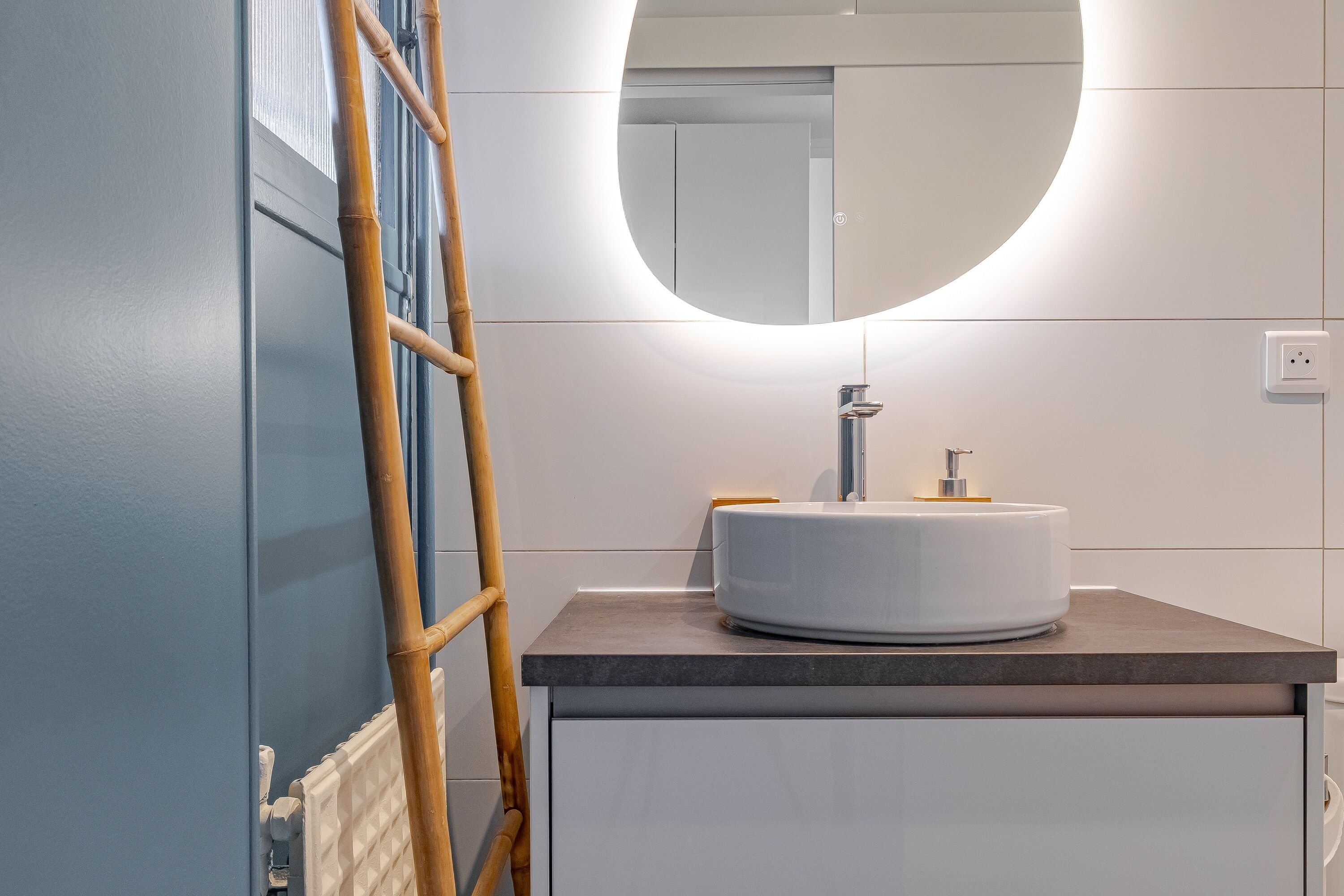 A bathroom with a round mirror, a white sink, a wooden ladder, and a white cabinet.