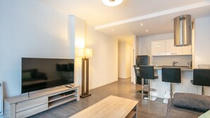Un salon moderne doté d’un téléviseur à écran plat, d’une table basse, d’un canapé, d’une lampe et d’un coin cuisine avec des tabourets de bar.