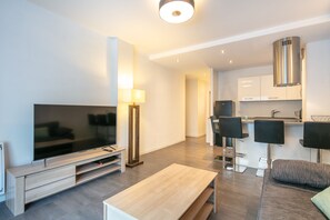 A modern living room with a flat-screen TV, a coffee table, a sofa, a lamp, and a kitchen area with bar stools. - Le Pradel Championnet - Bel appt centre Grenoble (Grenoble)