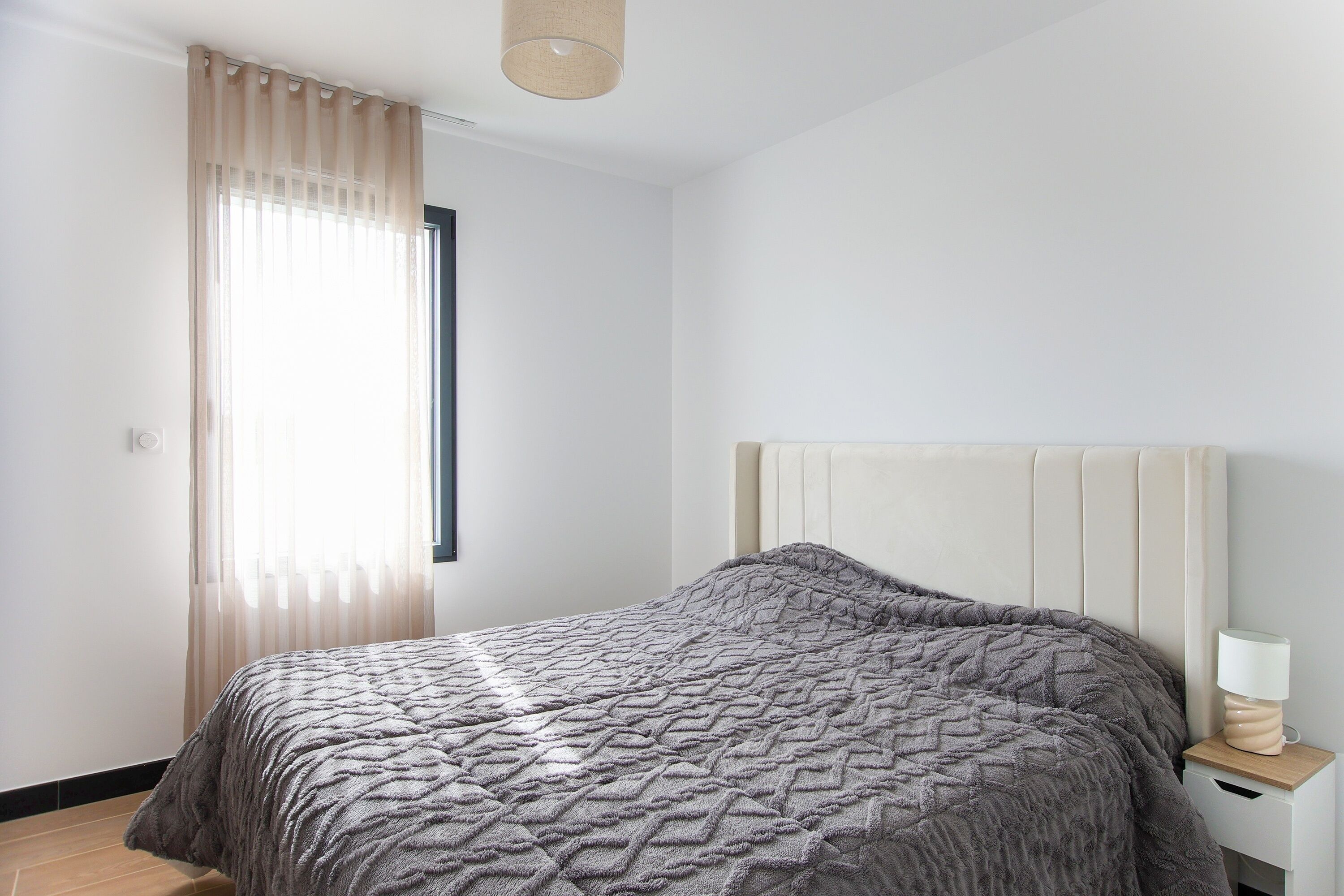 Ein Schlafzimmer mit einem Bett, einem Nachttisch mit Lampe, einem Fenster mit Vorhängen und einer Deckenleuchte.