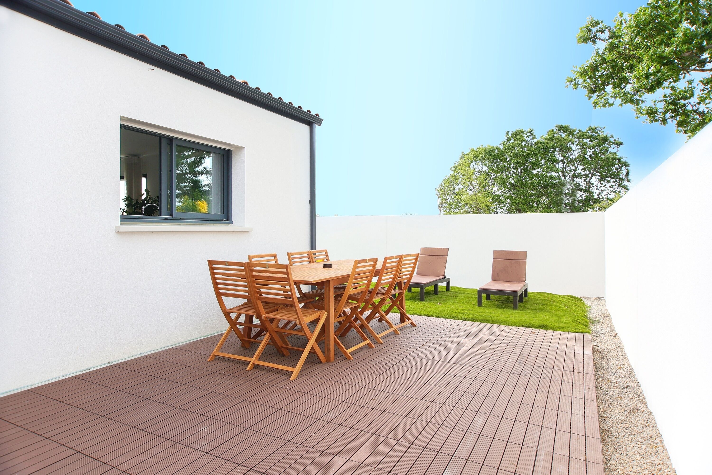 Eine Terrasse mit einem Holztisch und -stühlen, ein kleiner Gartenbereich und ein weißes Gebäude mit einem Fenster.