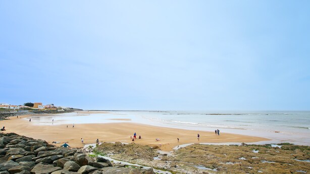 Una playa con gente, un primer plano rocoso y un edificio al fondo.