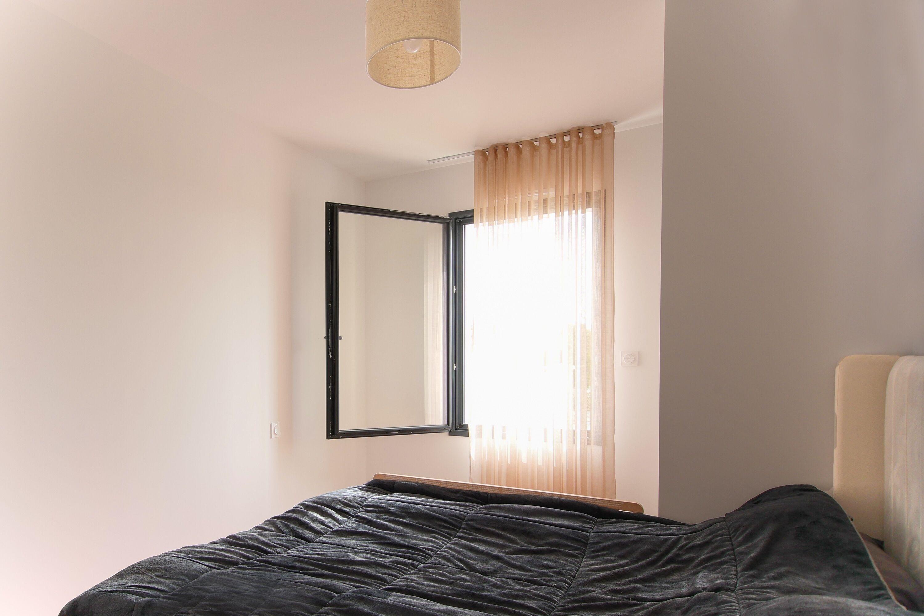 A bedroom with a bed, a window with curtains, a mirror, and a hanging light fixture.