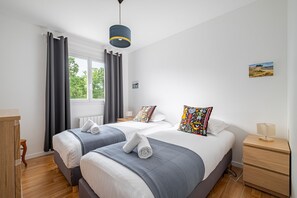 Ein Doppelbett mit grau-weißer Decke, ein Nachttisch mit Lampe, ein Fenster mit Vorhängen und ein kleines Bild an der Wand.