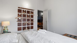 Un dormitorio con cama, mesita de noche con lámpara, un panel de madera con perchas en la pared, y una puerta que da a otra habitación.