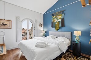 A bedroom with a bed, a nightstand, a lamp, a wall tapestry, and a door leading to an outdoor balcony. - Le Cocon de Béziers - Au cœur du centre-ville (Béziers)