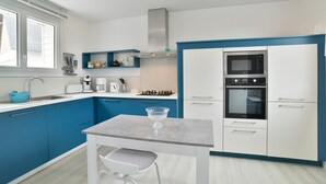 A modern kitchen with blue cabinets, a white countertop, a stainless steel range hood, a microwave, an oven, and a small dining table with a chair. - Les Grès roses - Appartement cosy sur la côte (Fréhel)