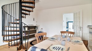 A dining area with a spiral staircase, wooden chairs, a striped tablecloth, and a view of the kitchen. - Les Grès roses - Appartement cosy sur la côte (Fréhel)