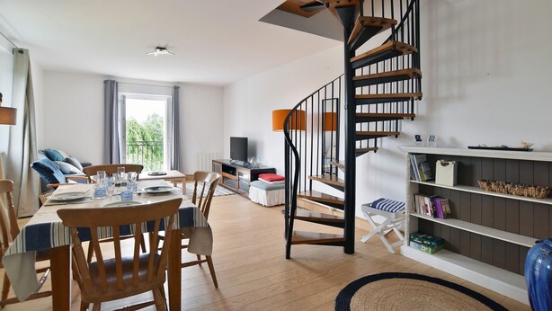 A modern living room with a wooden dining table, chairs, a staircase with a wooden railing, a bookshelf, and a television. - Les Grès roses - Appartement cosy sur la côte (Fréhel)