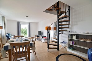 A modern living room with a wooden dining table, chairs, a staircase with a wooden railing, a bookshelf, and a television. - Les Grès roses - Appartement cosy sur la côte (Fréhel)
