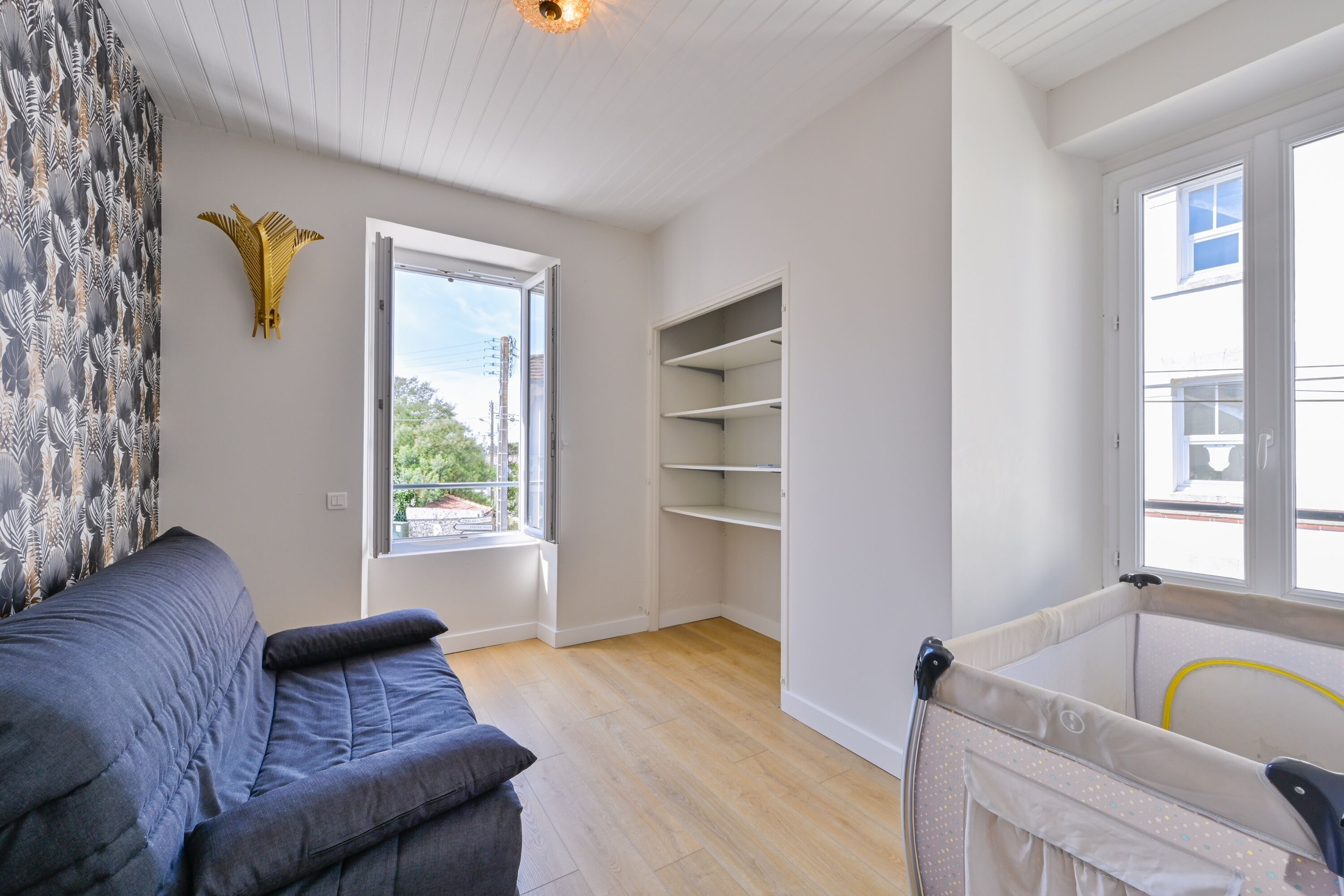A room with a grey sofa, a white shelving unit, a window with a view, and a white ceiling.