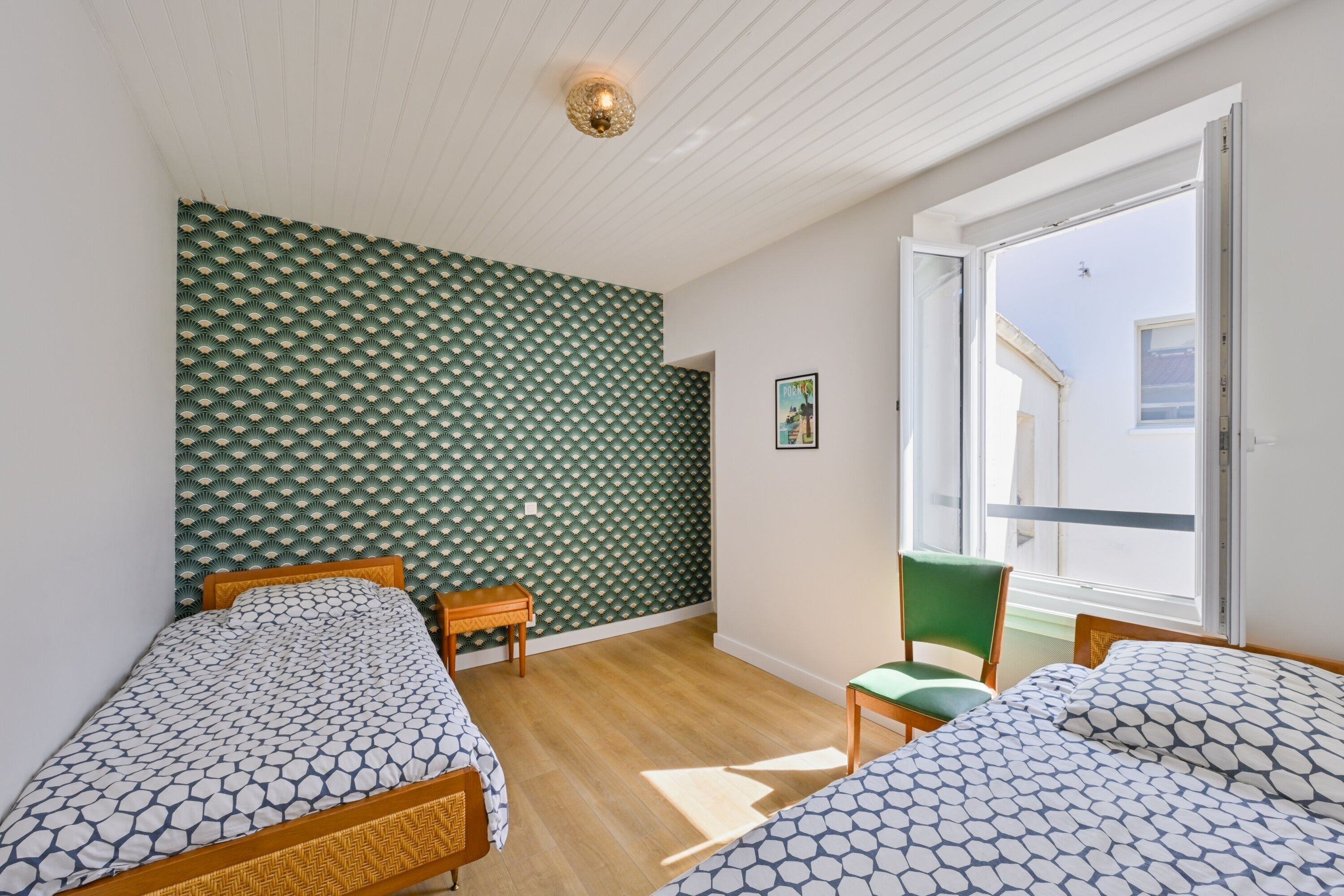 Une chambre avec deux lits, un plancher en bois, un mur à motifs verts, une fenêtre donnant sur un paysage agréable et une petite table.