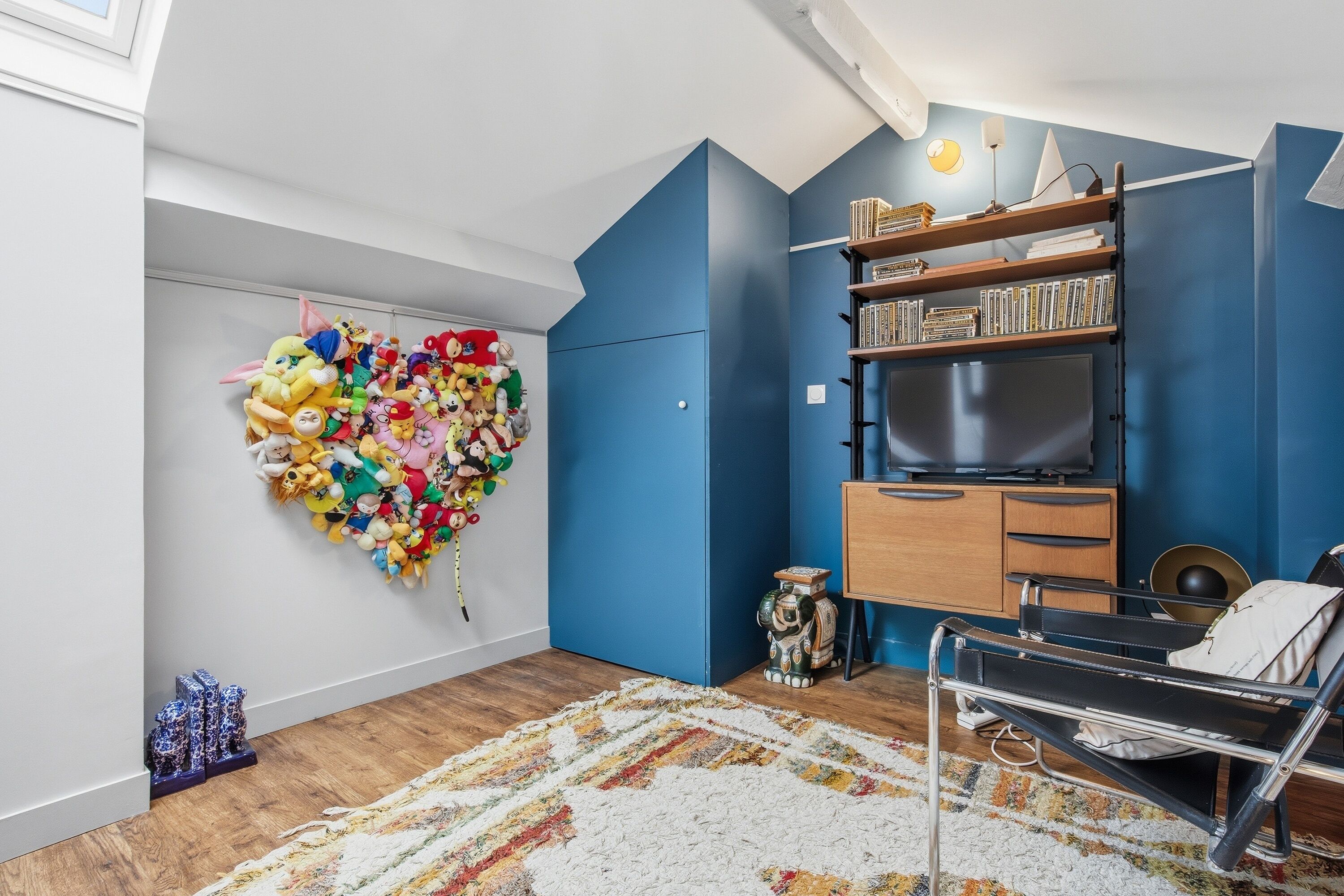 A room with a heart-shaped wall decoration of plush toys, a bookshelf, a sofa, and a patterned rug.