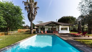 A rectangular swimming pool with a wooden deck, surrounded by a fenced yard, and a building with a tiled roof and large windows. - La Villa Amarra - À 1 km du Golf de Moliets-et-Maa (Moliets-et-Maa)