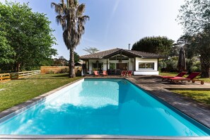 Ein rechteckiger Pool mit Holzdeck, umgeben von einem eingezäunten Garten und einem Gebäude mit geflastertem Dach und großen Fenstern.