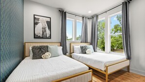 Ein Schlafzimmer mit zwei Betten, einem Fenster mit Vorhängen, einem Wandbild und einem gemusterten Vorhang links.
