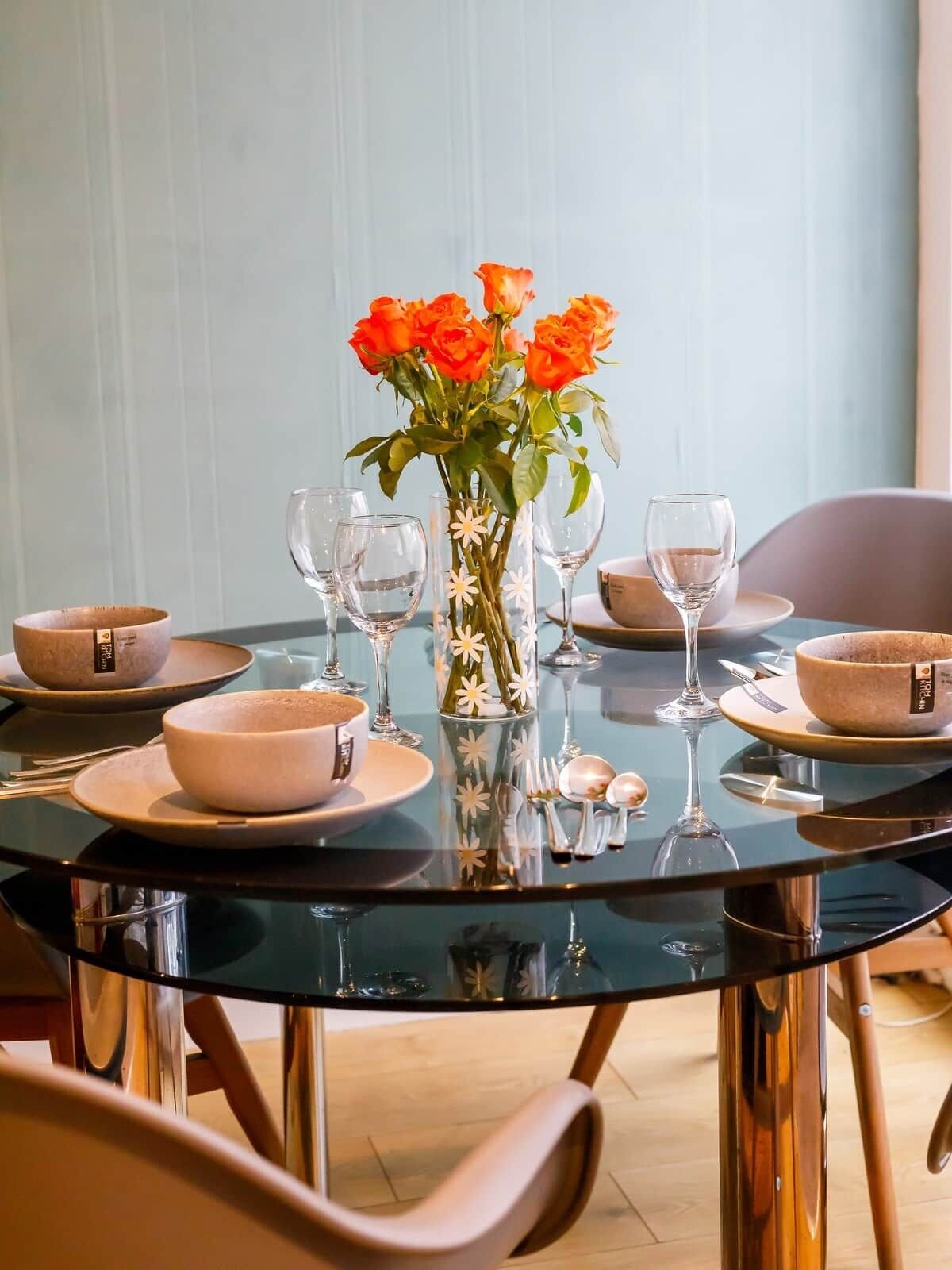 Uma mesa de jantar posta com pratos, tigelas e copos de vinho, acompanhada por um vaso de flores laranjas.
