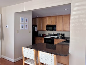 A compact kitchen with wooden cabinets, a microwave, a stove, and a sink. There are two white chairs with a woven backrest.
