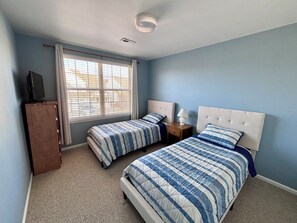 Un dormitorio con dos camas, un televisor sobre un mueble, una ventana con persianas y una lámpara en el techo.