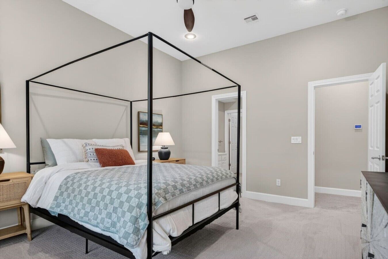 A four-poster bed with a checkered bedspread, two bedside tables with lamps, a wall-mounted painting, and a door leading to another room.