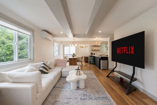 Moderni olohuone, jossa on valkoinen sohva, sohvapöytä ja televisio, jonka näytöllä näkyy teksti ’NETFLIX’.