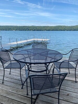 A round metal table with chairs on a wooden deck by a lake. - Owasco Lake Cottage | Lake Front | Dock | Fire Pit (Moravia)