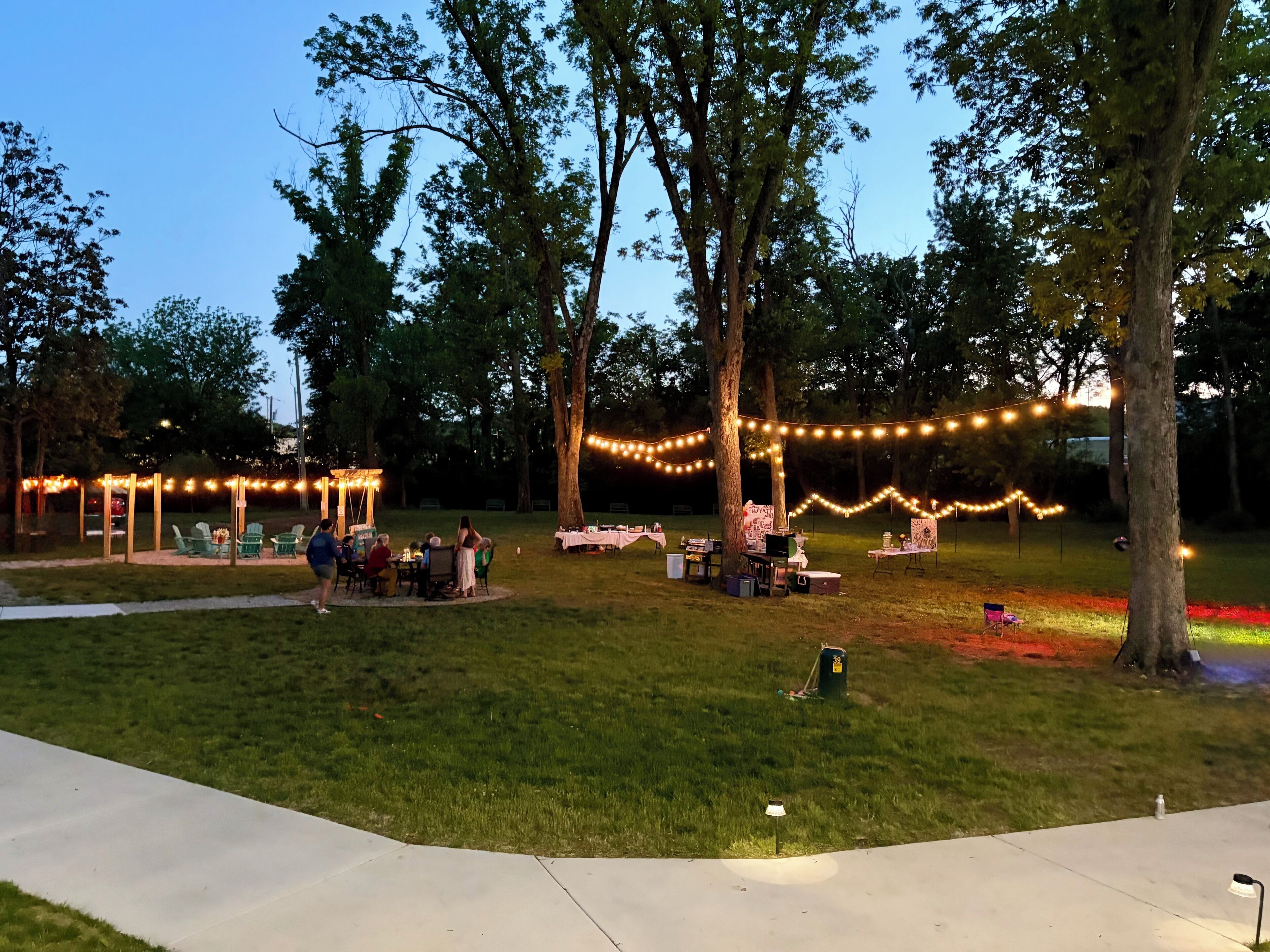 An outdoor event area with tables and chairs, string lights, and trees.