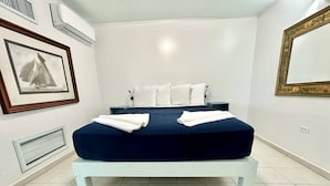 A neatly made bed with white pillows and a dark blue bedspread, a framed picture on the wall, and a white air conditioning unit. - Apto 2A Flamenco Beachfront Villas with Ocean View (Flamenco)