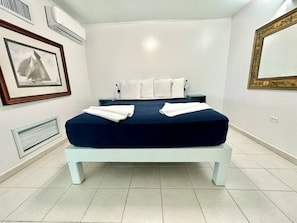 Un lit bien fait, avec des oreillers blancs et une couvre-lit bleu foncé, un tableau encadré au mur et un appareil de climatisation blanc.