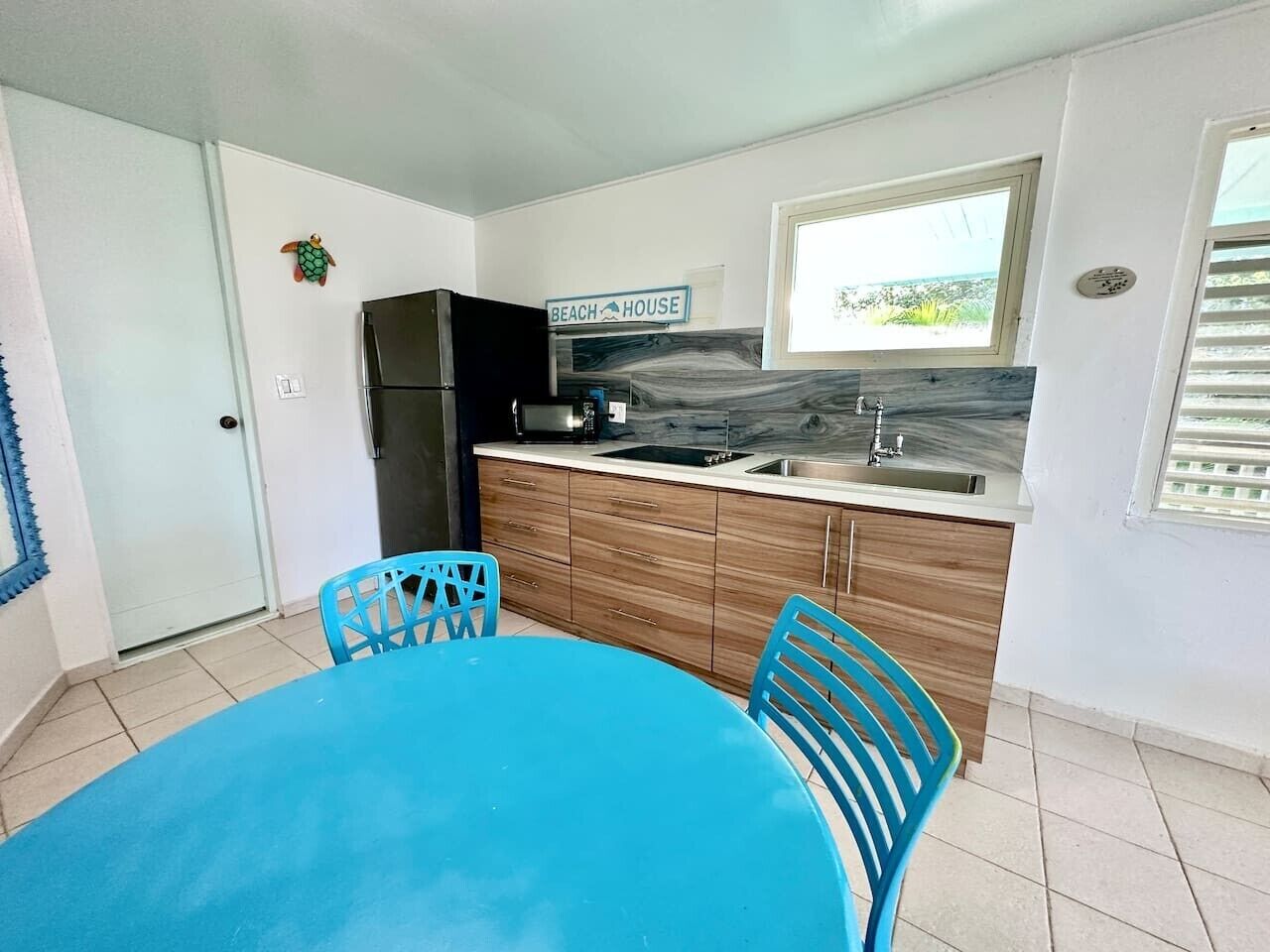 Una cucina con tavolo e sedie azzurre, un frigorifero, pensili in legno, un cartello con su scritto 'BEACH HOUSE' e una finestra vista mare.