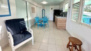 A room with a white chair, a small wooden stool, a blue table with chairs, a kitchenette, and a door with a glass panel. - Apto 2A Flamenco Beachfront Villas with Ocean View (Flamenco)