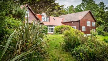A large, two-story wooden house with a sloped roof, surrounded by lush greenery and trees.