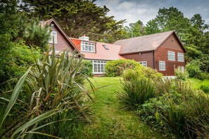 A large, two-story wooden house with a sloped roof, surrounded by lush greenery and trees.