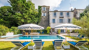 A pool area with lounge chairs and umbrellas, surrounded by greenery and a stone building. - Le Jardin Du Chateau (Duras)